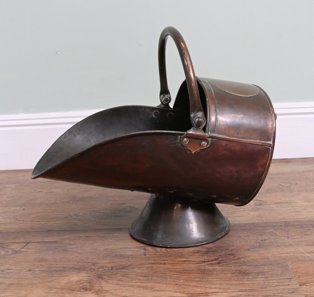 A Victorian copper helmet-shaped coal scuttle featuring a flared pedestal base and an arched swing handle.