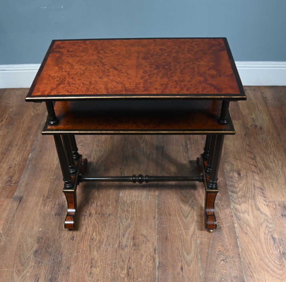 A late Victorian two-tier dumb waiter with warm amboyna wood surfaces, ebonised turned column supports, and splayed legs on castors. View II