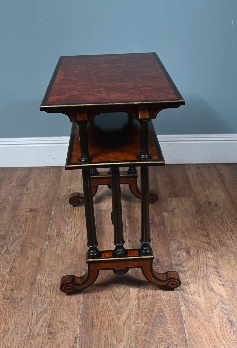 A late Victorian two-tier dumb waiter with warm amboyna wood surfaces, ebonised turned column supports, and splayed legs on castors. View V