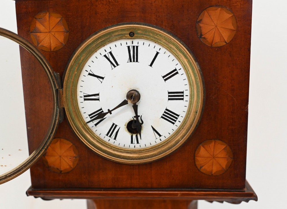 A miniature mahogany longcase clock featuring classical Sheraton style wood inlay with an urn motif and a circular white dial with Roman numerals. View IV