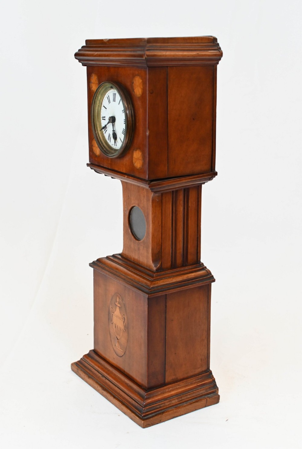 A miniature mahogany longcase clock featuring classical Sheraton style wood inlay with an urn motif and a circular white dial with Roman numerals. View VI