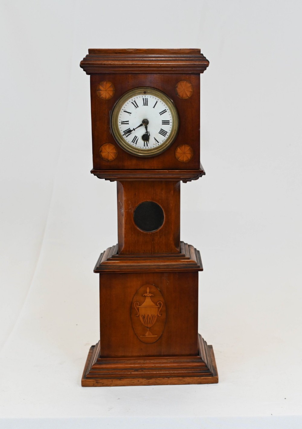 A miniature mahogany longcase clock featuring classical Sheraton style wood inlay with an urn motif and a circular white dial with Roman numerals. View VII