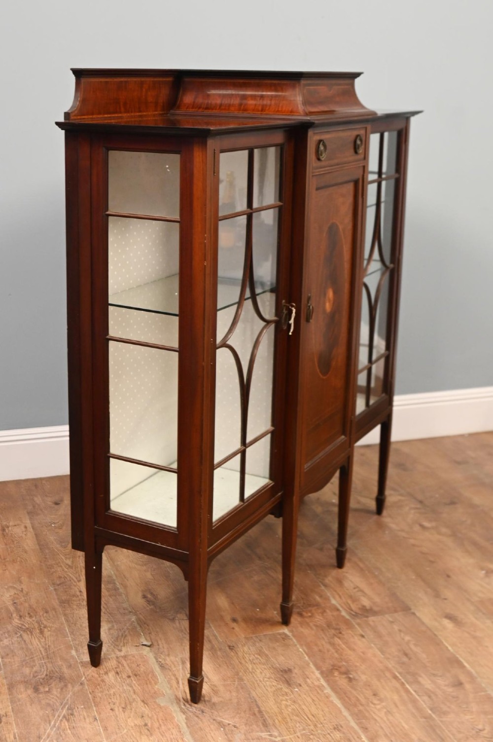 An Edwardian inlaid mahogany display cabinet featuring astragal glazed side doors, a central wooden cupboard with oval marquetry, and slender square tapered legs. View II