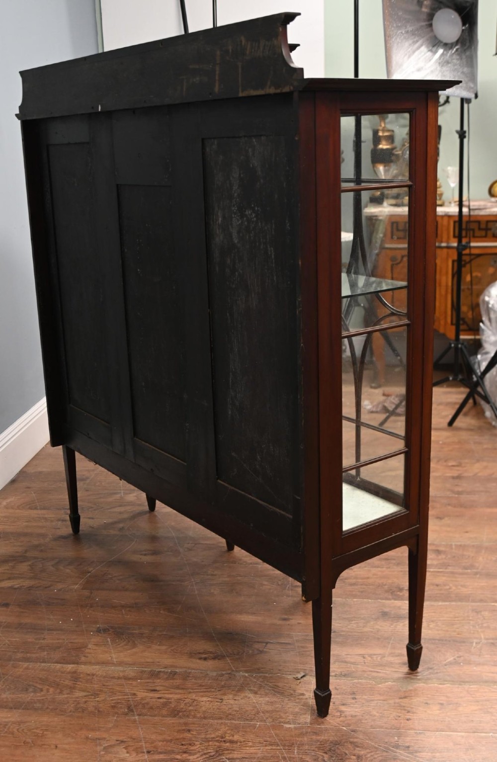 An Edwardian inlaid mahogany display cabinet featuring astragal glazed side doors, a central wooden cupboard with oval marquetry, and slender square tapered legs. View IV