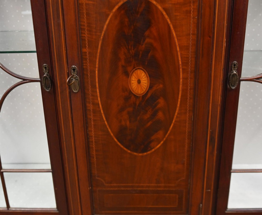 An Edwardian inlaid mahogany display cabinet featuring astragal glazed side doors, a central wooden cupboard with oval marquetry, and slender square tapered legs. View VI