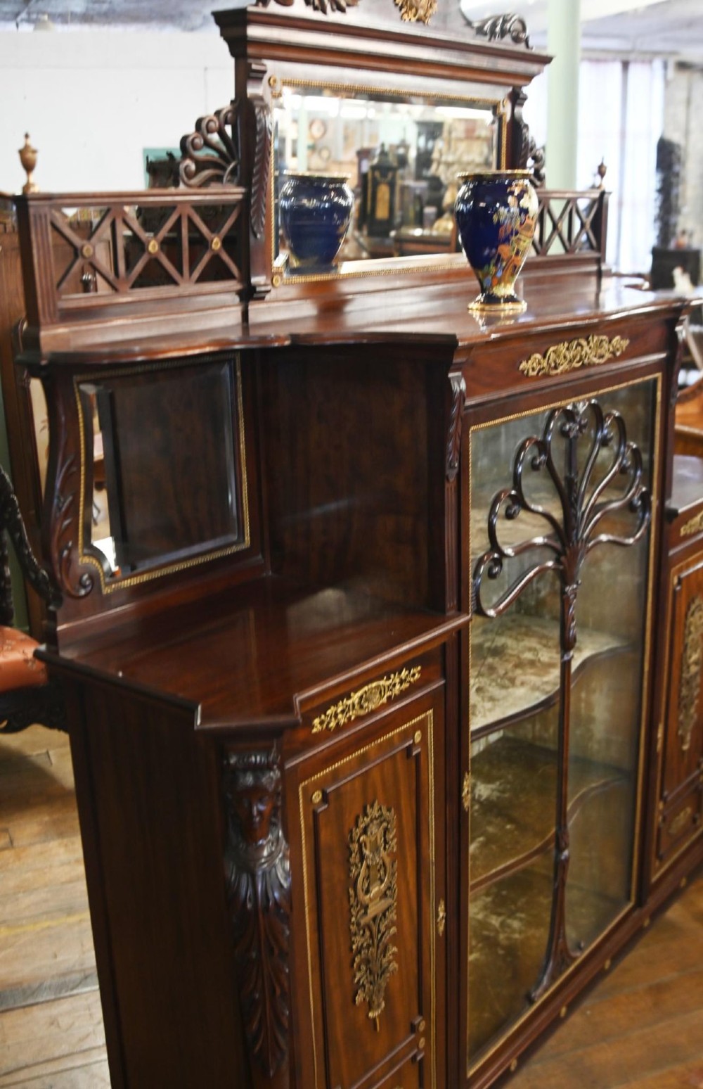 A dark mahogany display cabinet featuring gilt ormolu mounts, bevelled mirror panels, and a central glass door with decorative organic glazing bars. View III