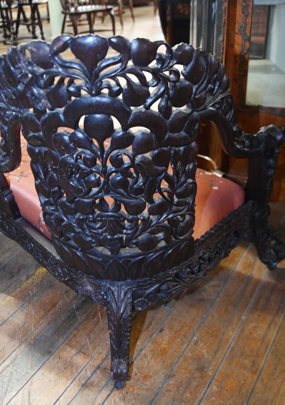An intricately carved 19th-century dark teak sofa featuring openwork floral motifs and a terracotta floral upholstered seat pad. View IV