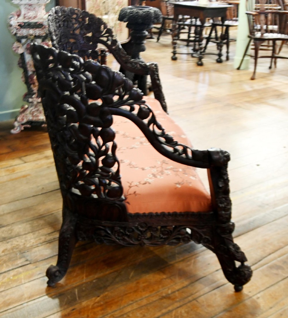 An intricately carved 19th-century dark teak sofa featuring openwork floral motifs and a terracotta floral upholstered seat pad. View V