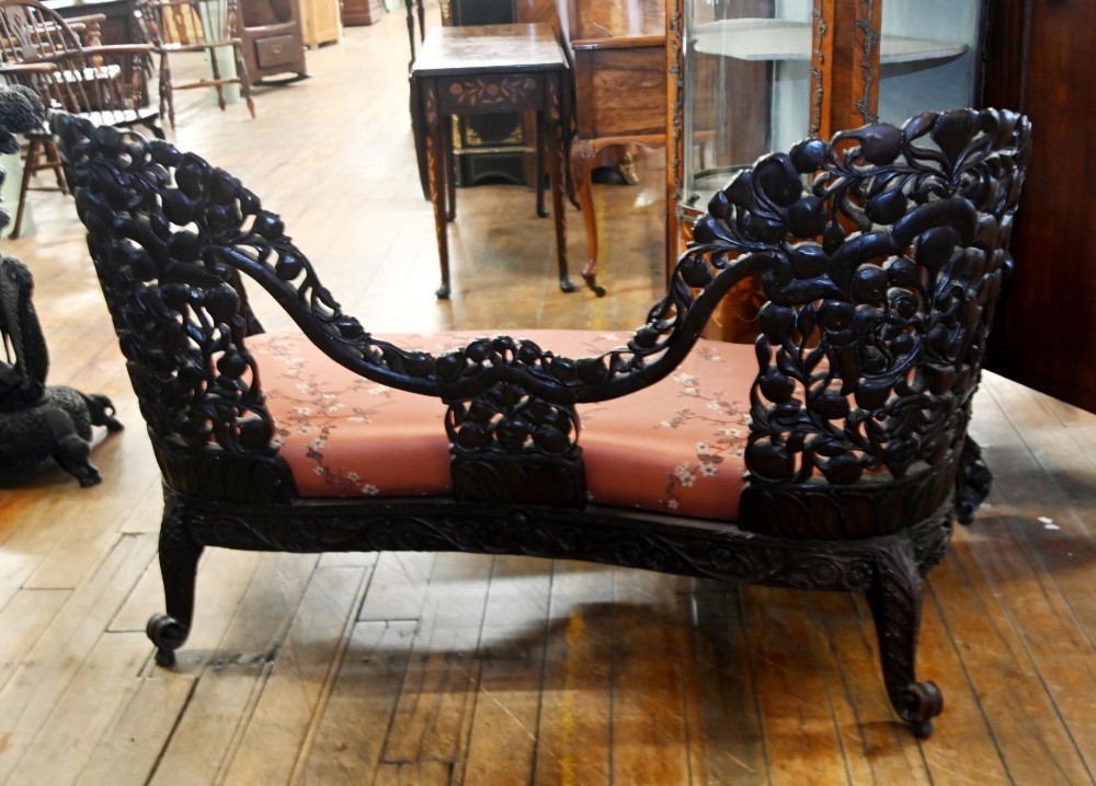 An intricately carved 19th-century dark teak sofa featuring openwork floral motifs and a terracotta floral upholstered seat pad. View VI