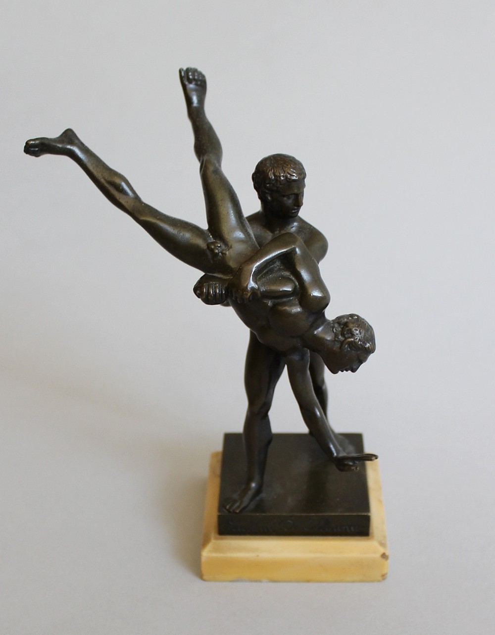 A dark bronze sculpture depicting two classical male wrestlers engaged in combat, mounted on a light marble base.