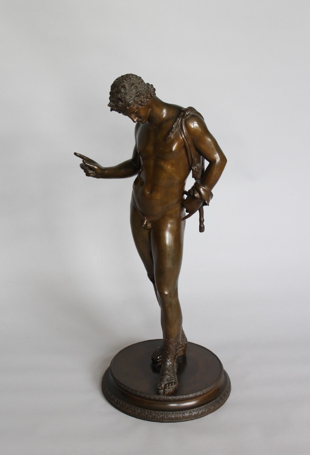A dark brown patinated bronze statue depicting the classical male figure of Narcissus standing on a circular decorative base.