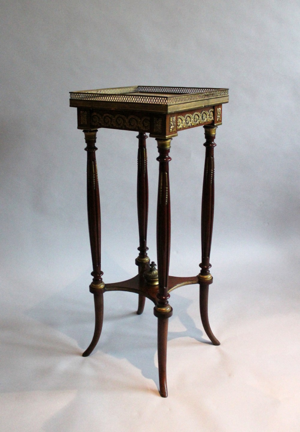 A tall, slender dark mahogany French occasional table featuring a pierced brass gallery top, fluted legs, and a low curved x-stretcher with a brass finial.