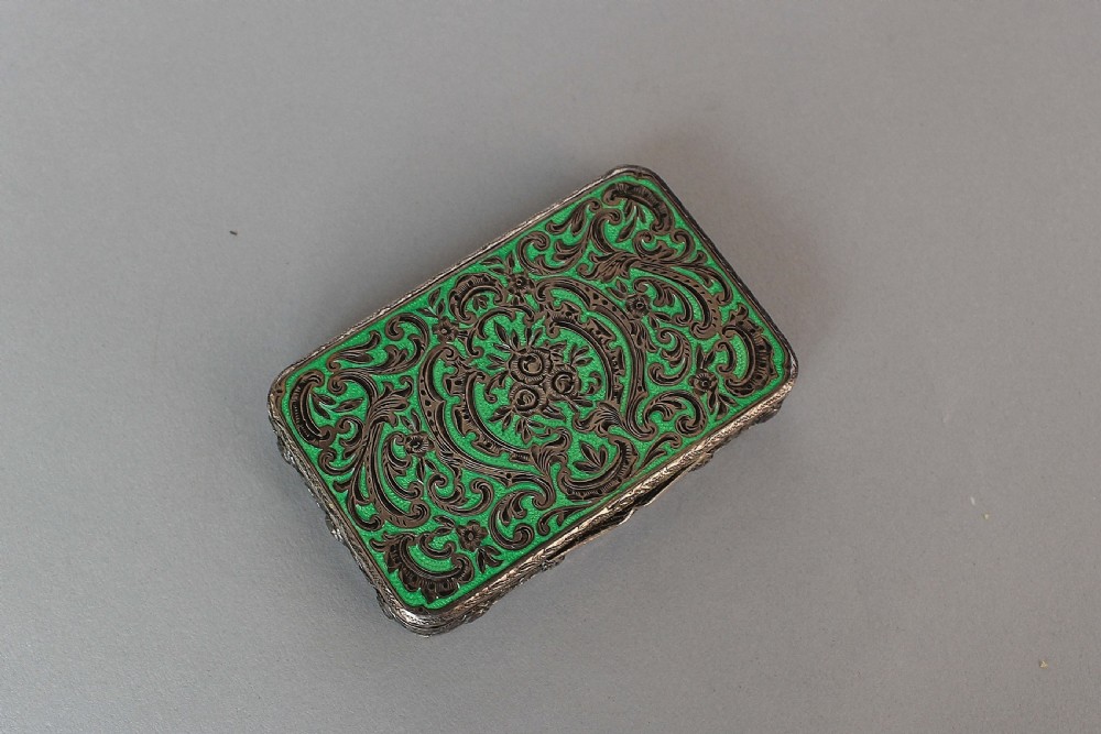 A small rectangular silver box featuring ornate foliate scrollwork overlaid upon a vibrant green engine-turned enamel surface.