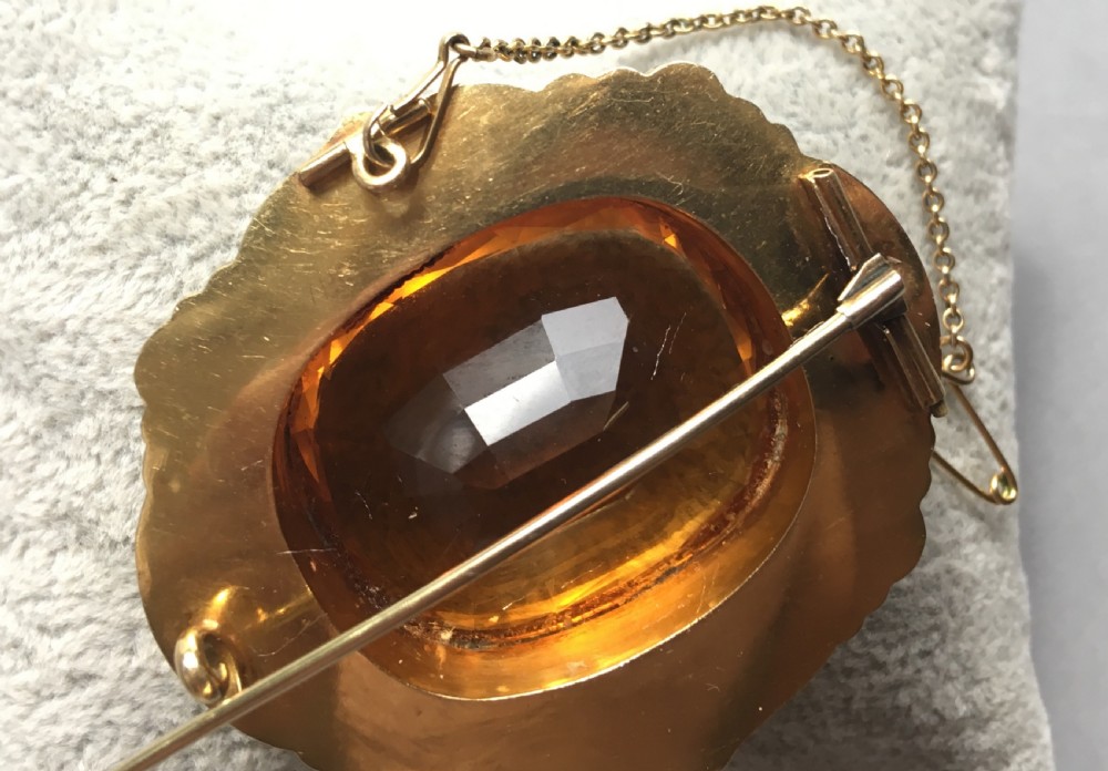 An antique yellow gold brooch set with a large faceted oval citrine gemstone surrounded by an ornate scrollwork frame with an attached safety chain. View II