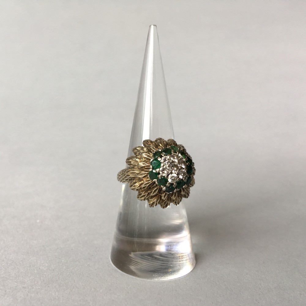 A textured yellow gold cocktail ring featuring a central cluster of round brilliant diamonds surrounded by emeralds and feathered metalwork.