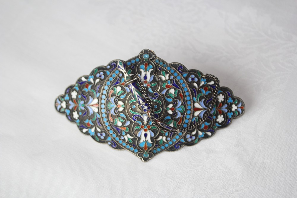 A lobed silver belt buckle decorated with intricate polychrome floral cloisonne enamel in shades of turquoise, navy, white, and green, complete with a small dagger-shaped fastening pin.