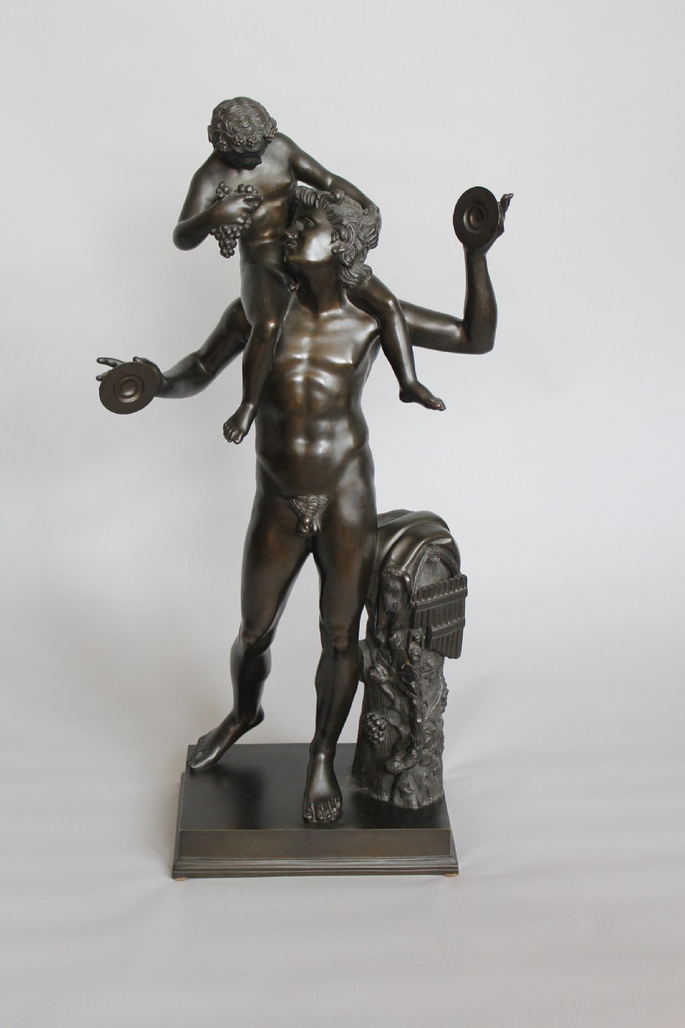 A dark patinated bronze sculpture depicting a male faun holding cymbals with the infant Bacchus seated on his shoulders, mounted on a rectangular plinth base.