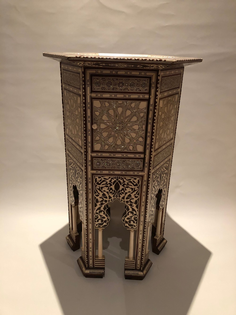 An octagonal dark wood side table with intricate bone and mother-of-pearl geometric inlay, featuring carved horseshoe arches and turned legs at the base.