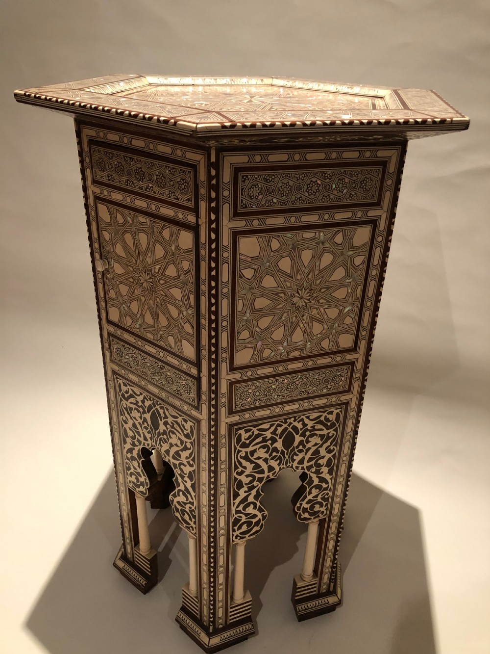 An octagonal dark wood side table with intricate bone and mother-of-pearl geometric inlay, featuring carved horseshoe arches and turned legs at the base. View V