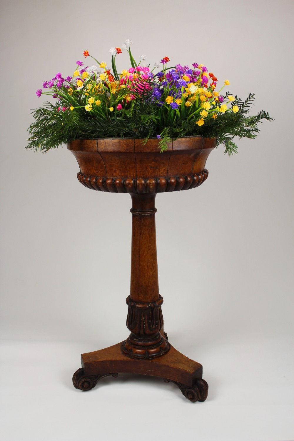 A golden oak jardiniere stand featuring a carved circular basin, a turned central column, and a triform base with scroll feet.