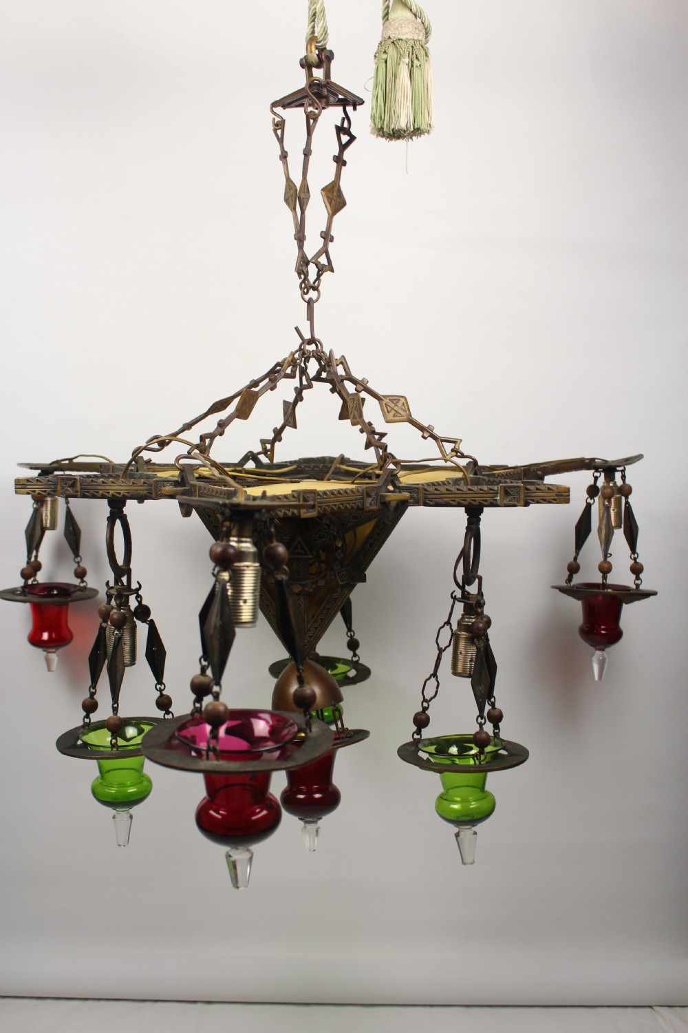 An Arts and Crafts style brass seven-light chandelier featuring geometric framework and suspended red and green glass cups.