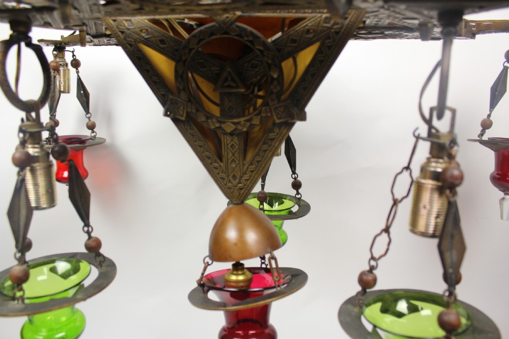 An Arts and Crafts style brass seven-light chandelier featuring geometric framework and suspended red and green glass cups. View II