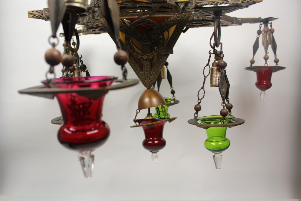An Arts and Crafts style brass seven-light chandelier featuring geometric framework and suspended red and green glass cups. View III