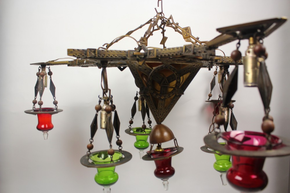 An Arts and Crafts style brass seven-light chandelier featuring geometric framework and suspended red and green glass cups. View IV