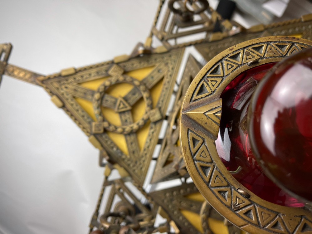 An Arts and Crafts style brass seven-light chandelier featuring geometric framework and suspended red and green glass cups. View V