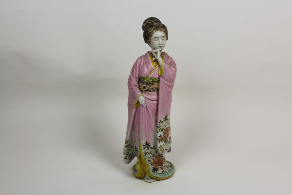 A hand-painted porcelain figurine of a Japanese geisha wearing a pink floral kimono with hand resting near her chin. View II
