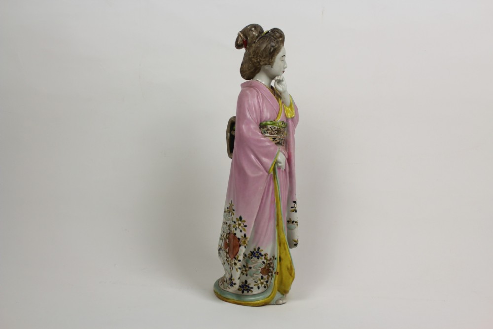 A hand-painted porcelain figurine of a Japanese geisha wearing a pink floral kimono with hand resting near her chin. View IV