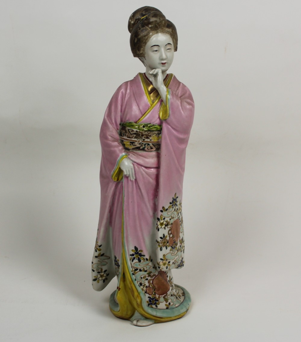 A hand-painted porcelain figurine of a Japanese geisha wearing a pink floral kimono with hand resting near her chin.