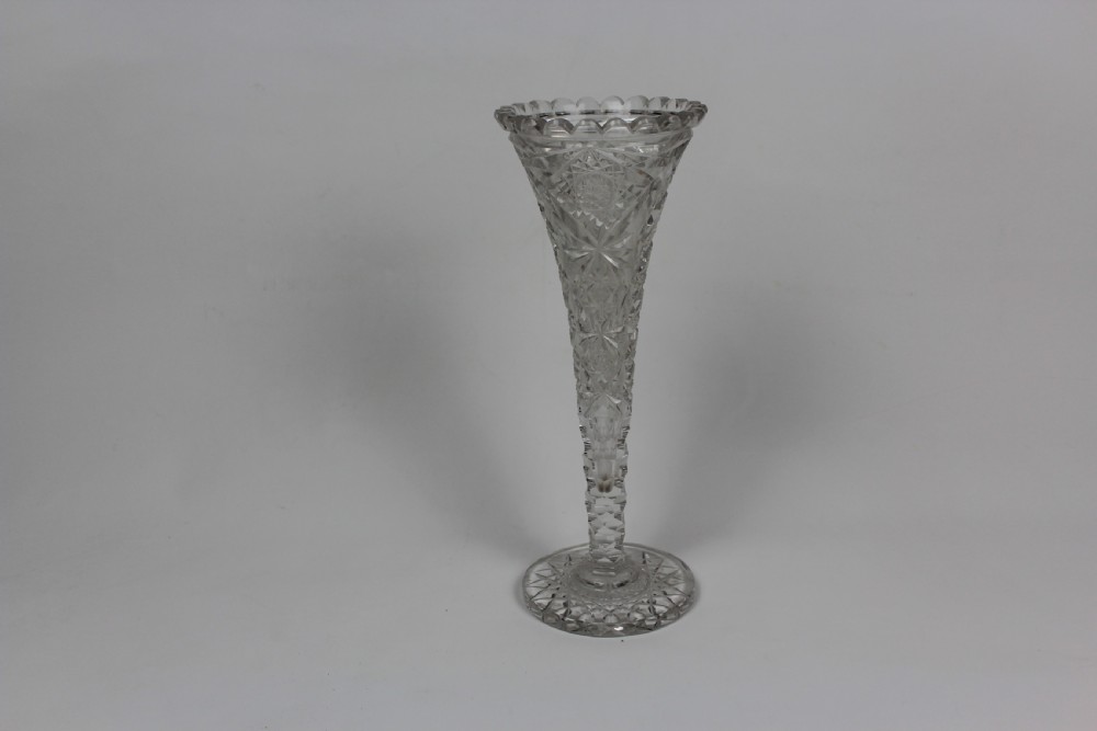 A tall trumpet-shaped clear hand-cut crystal vase featuring diamond cuts, a scalloped top rim, and a circular starburst pattern base.