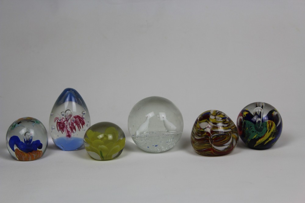 A collection of six assorted glass paperweights featuring various internal floral motifs, colourful swirls, and controlled bubbles.