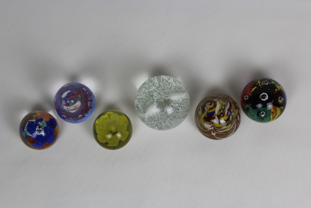 A collection of six assorted glass paperweights featuring various internal floral motifs, colourful swirls, and controlled bubbles. View IV