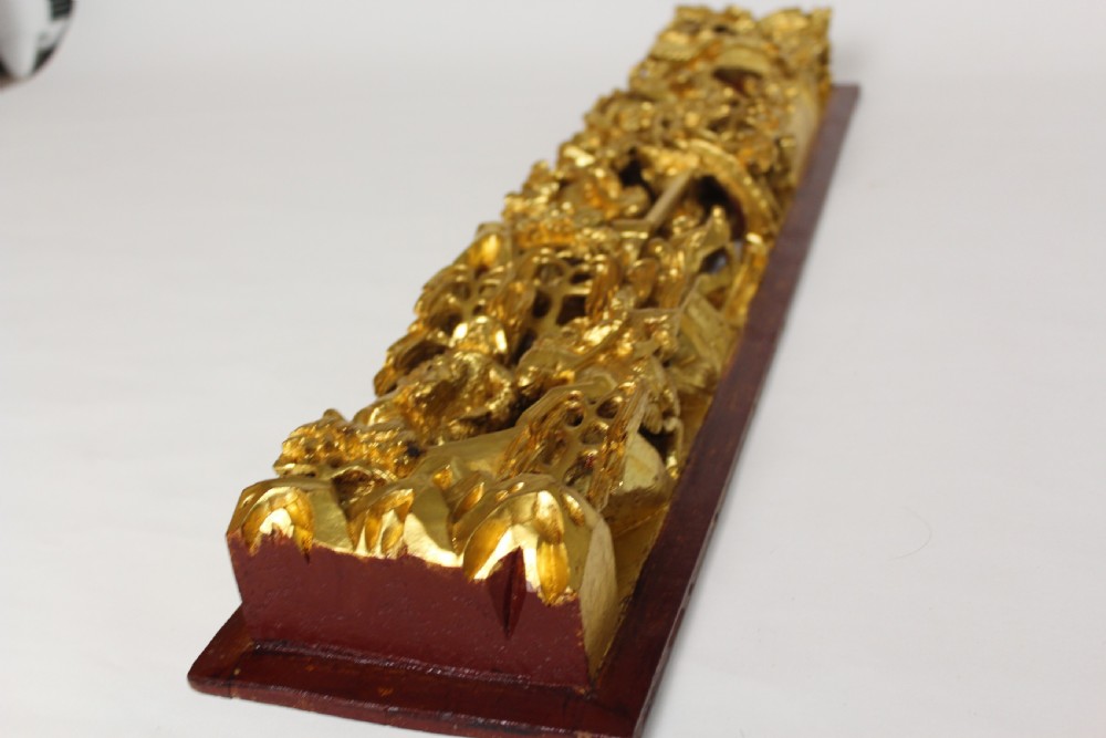 An elongated rectangular Chinese wooden wall plaque featuring deeply carved figures coated in gold leaf mounted on a red backing.