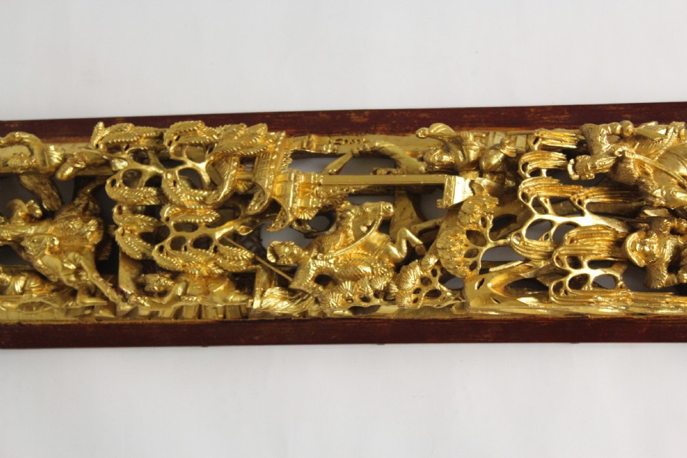 An elongated rectangular Chinese wooden wall plaque featuring deeply carved figures coated in gold leaf mounted on a red backing. View II