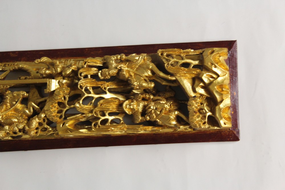 An elongated rectangular Chinese wooden wall plaque featuring deeply carved figures coated in gold leaf mounted on a red backing. View III