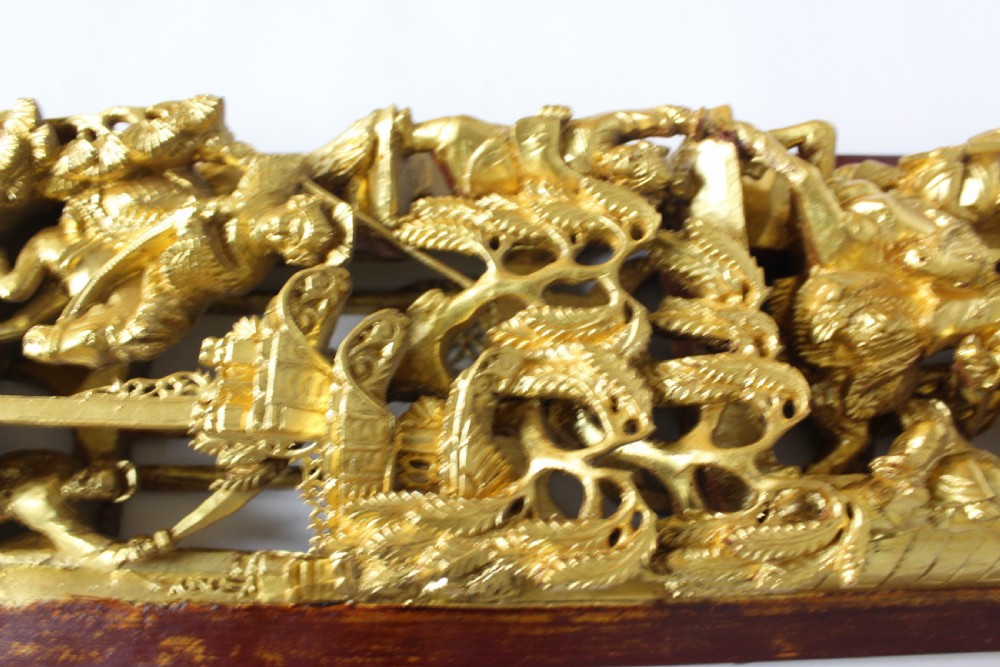 An elongated rectangular Chinese wooden wall plaque featuring deeply carved figures coated in gold leaf mounted on a red backing. View IV