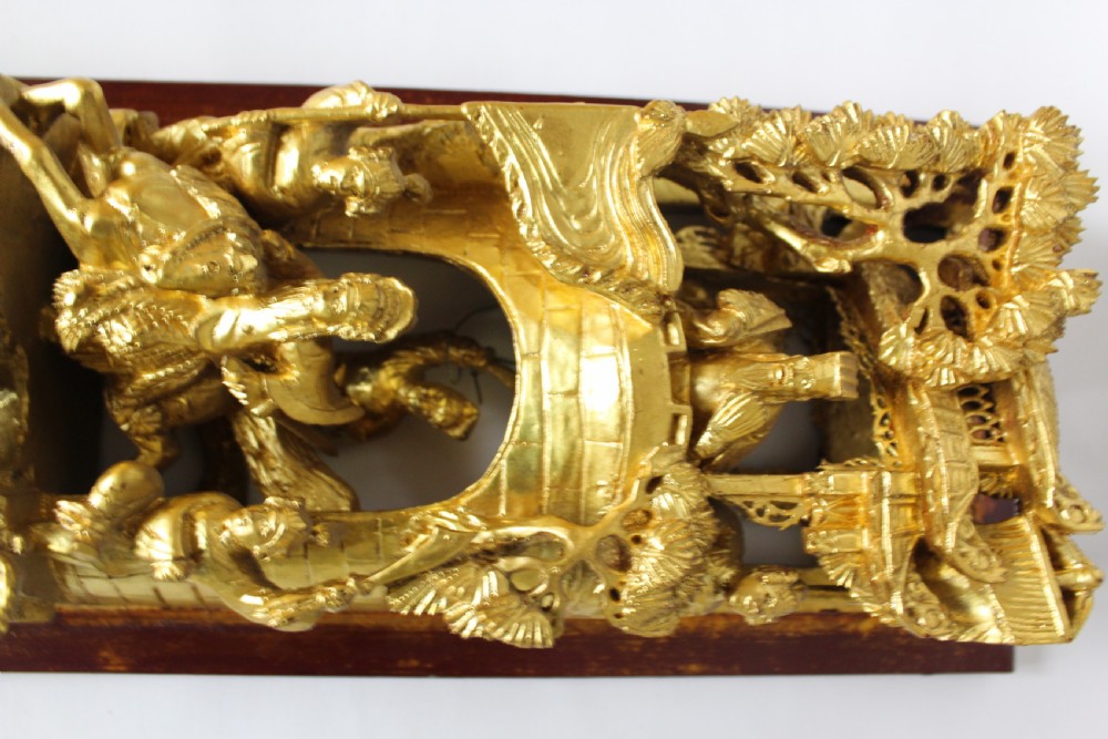 An elongated rectangular Chinese wooden wall plaque featuring deeply carved figures coated in gold leaf mounted on a red backing. View V