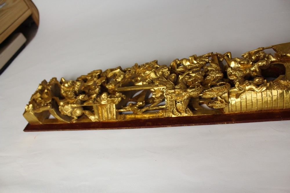 An elongated rectangular Chinese wooden wall plaque featuring deeply carved figures coated in gold leaf mounted on a red backing. View VI