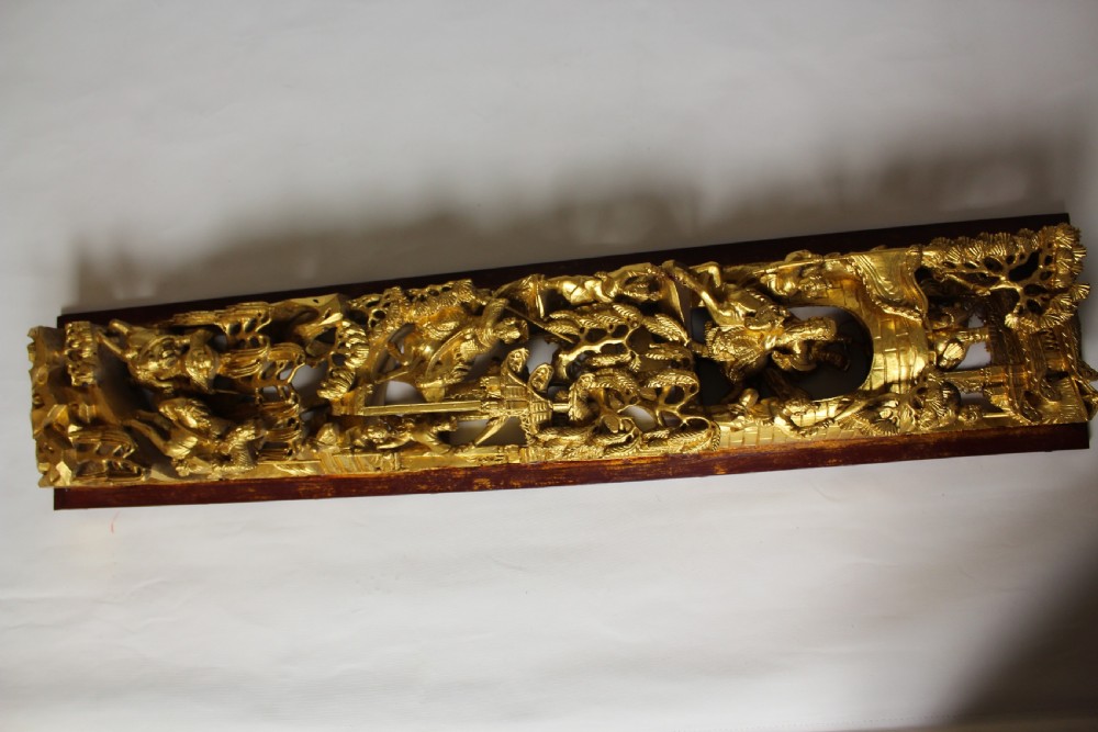 An elongated rectangular Chinese wooden wall plaque featuring deeply carved figures coated in gold leaf mounted on a red backing. View VII