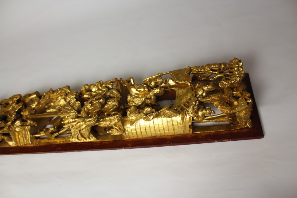 An elongated rectangular Chinese wooden wall plaque featuring deeply carved figures coated in gold leaf mounted on a red backing. View VIII