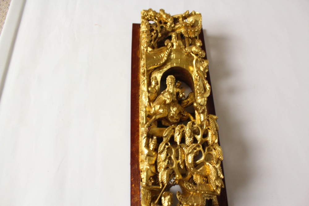 An elongated rectangular Chinese wooden wall plaque featuring deeply carved figures coated in gold leaf mounted on a red backing. View IX