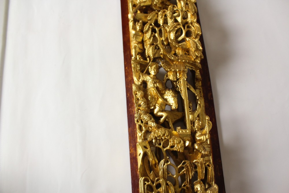 An elongated rectangular Chinese wooden wall plaque featuring deeply carved figures coated in gold leaf mounted on a red backing. View X
