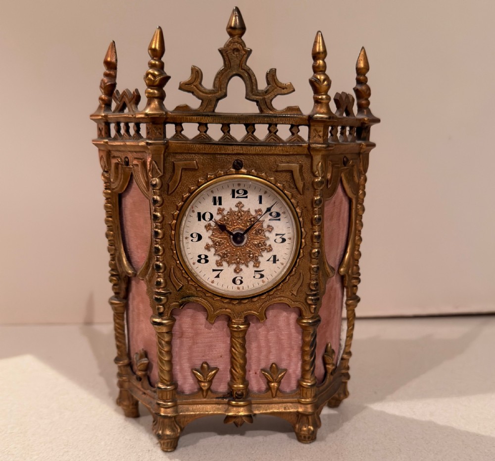 An ornate Victorian Gothic Revival gilt brass desk clock featuring pointed finials, twisted columns, and pleated pink silk side panels surrounding a central circular dial.