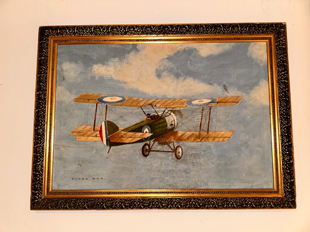A framed oil painting depicting a green vintage biplane flying against a clouded blue sky, signed by Simon Orr.