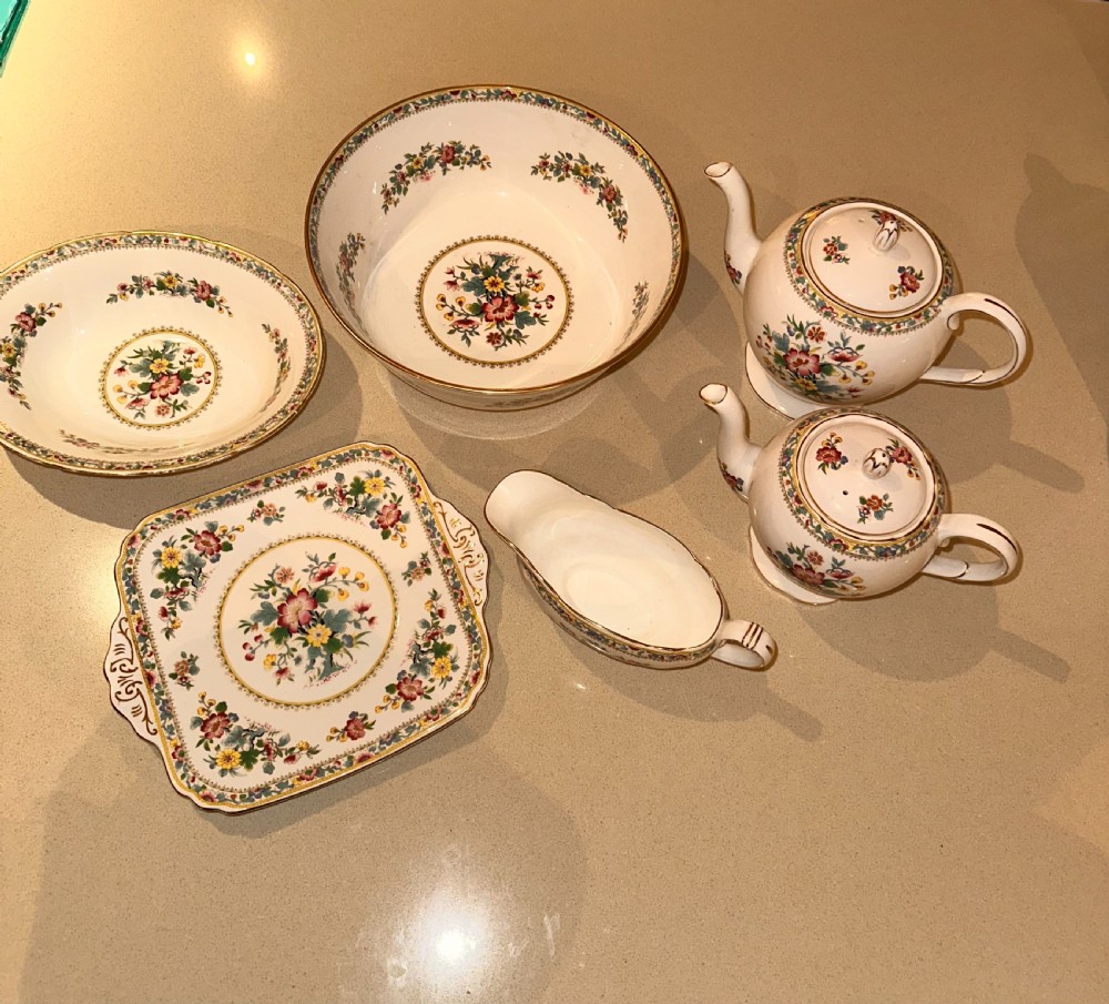 A collection of cream ceramic tableware featuring hand-painted floral motifs and gilded rims, including two teapots, a serving bowl, a gravy boat, and a square platter.
