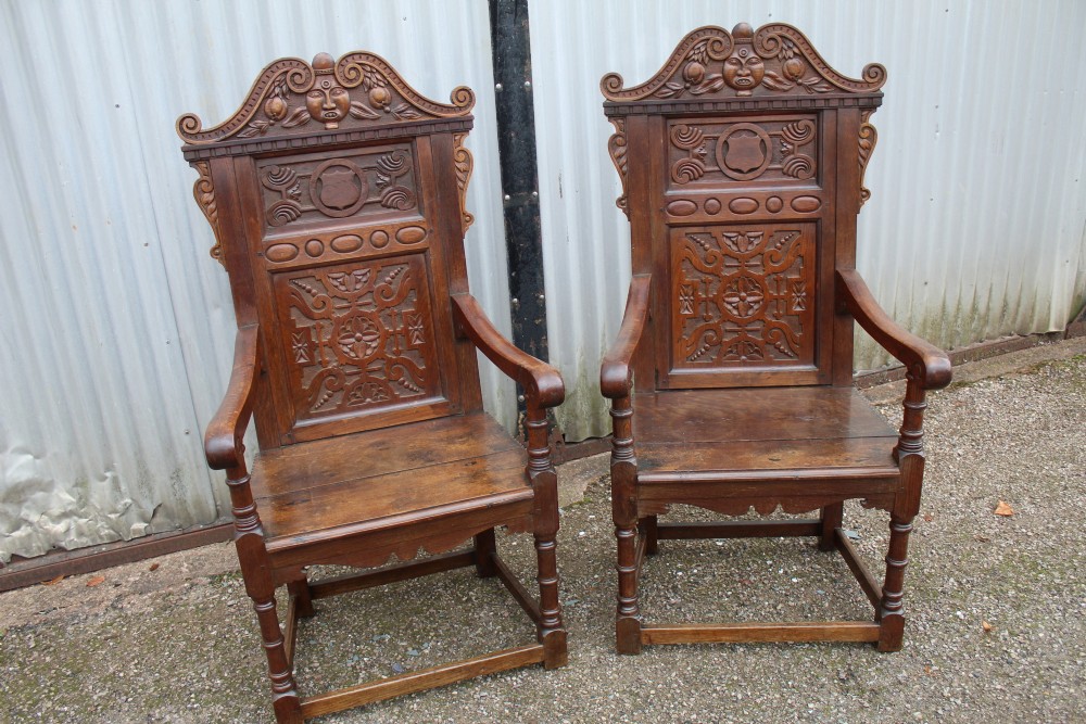 A pair of carved dark oak wainscot armchairs with relief-panelled backs, turned front legs, and flat armrests.