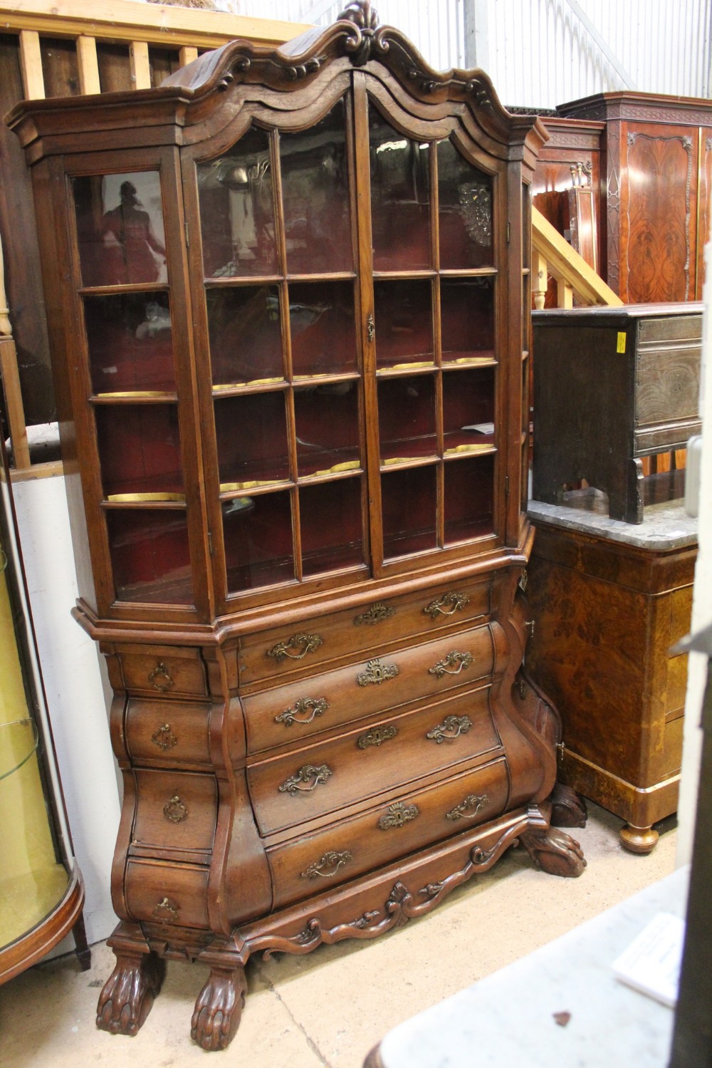 A Dutch Rococo walnut vitrine display cabinet featuring a shaped carved pediment, multi-pane glazed doors, and a four-drawer bombe chest base resting on carved claw feet.